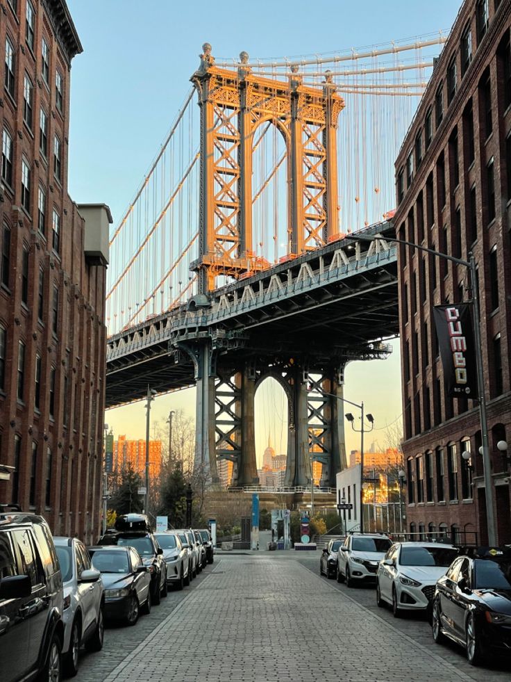 cars are parked in front of buildings and the bridge is high above them, as seen from an alleyway