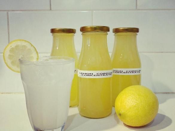 three lemonade bottles next to a glass filled with liquid and two lemons on the counter