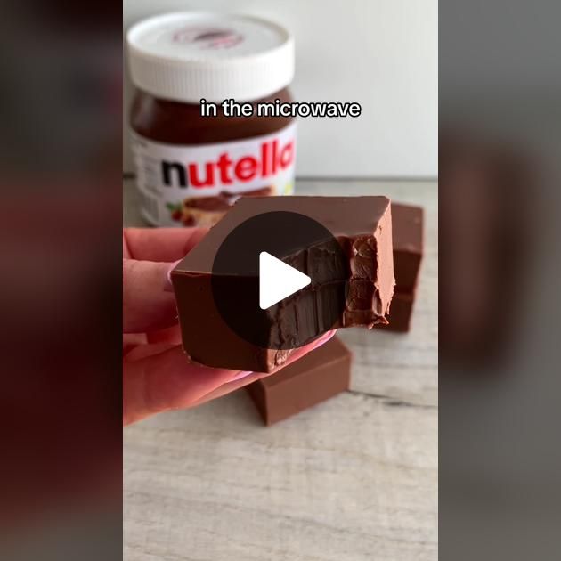 a hand holding a piece of chocolate with nutella in the background and a jar of nutella next to it