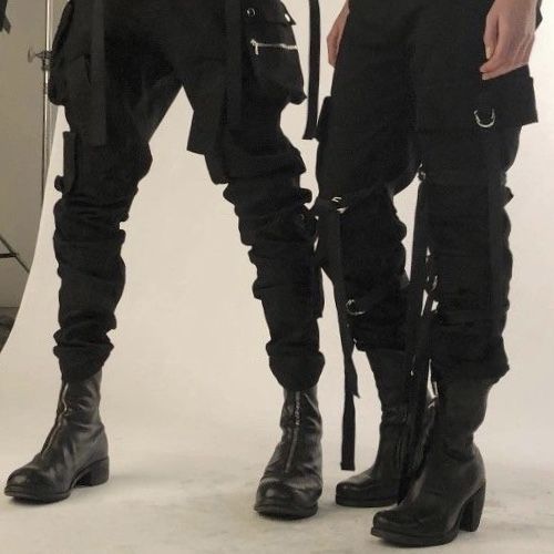 two men standing next to each other in front of a white background wearing black pants and boots