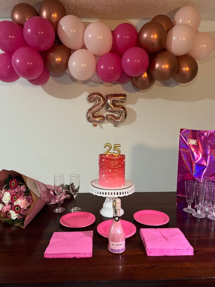 a table topped with a cake and pink napkins next to balloons on the wall