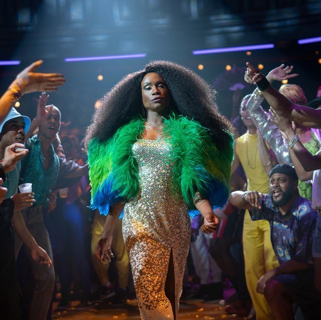 a woman in a green and blue feathered jacket is dancing on stage with other people