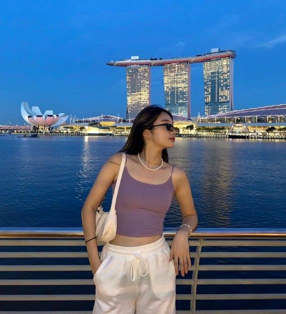 a woman standing next to the water in front of some tall buildings with lights on them