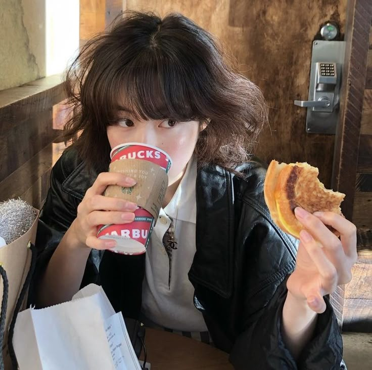 a woman sitting at a table drinking from a cup and holding a donut in her hand