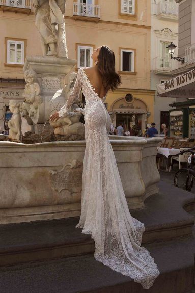 a woman standing in front of a fountain wearing a long sleeved wedding dress with open back