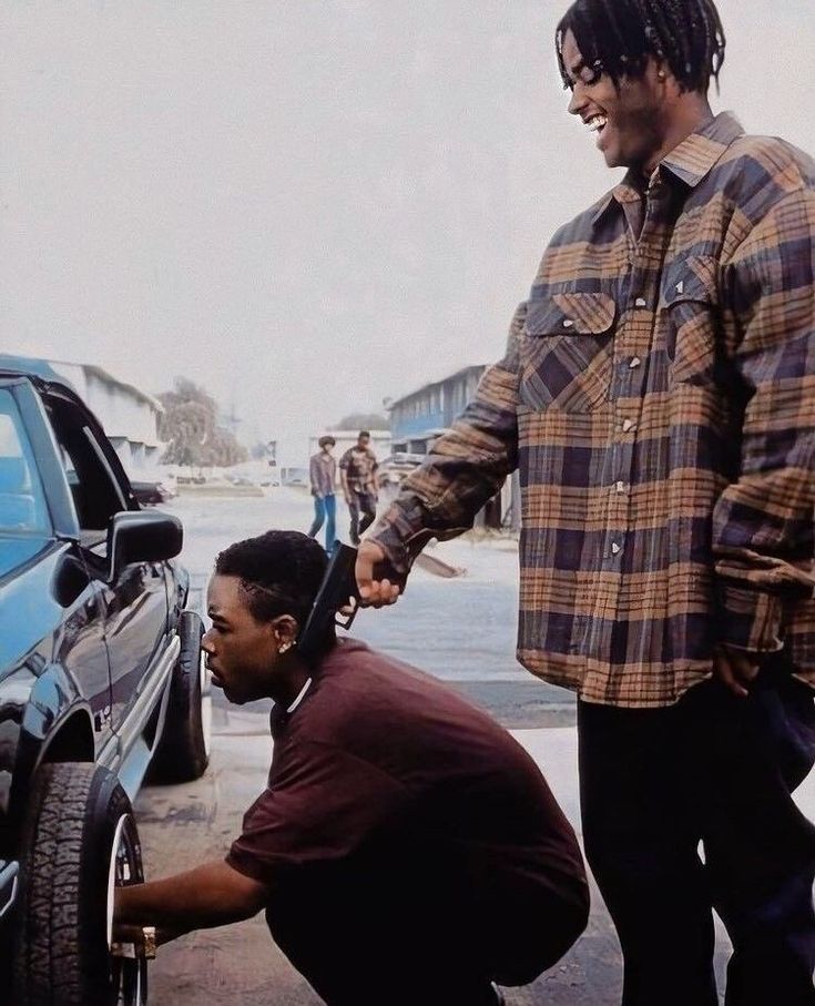 two men standing next to each other in front of a car and one is fixing the tire