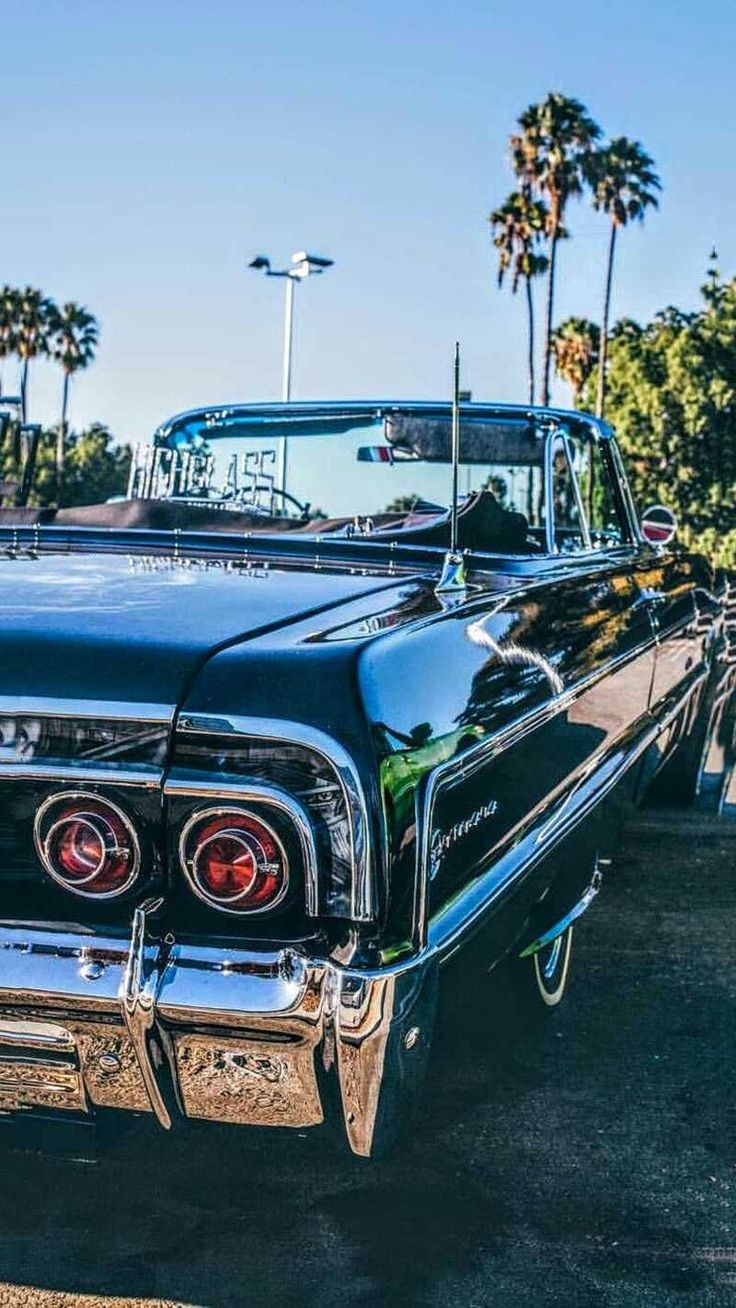 an old black car is parked in a parking lot with palm trees and other cars