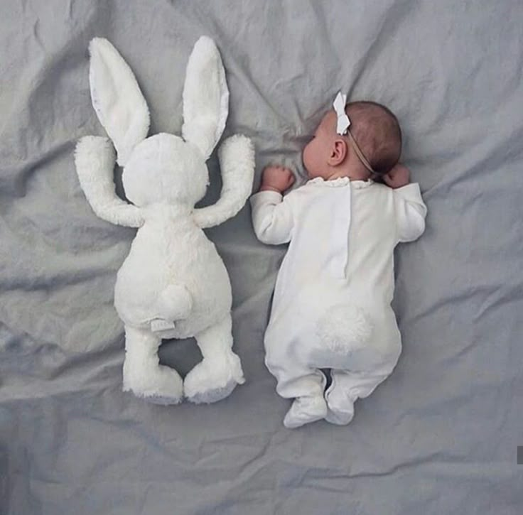 a baby laying next to a stuffed rabbit on top of a white sheet covered bed