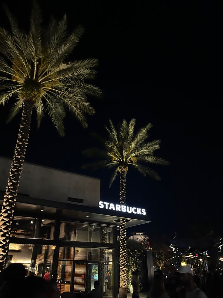 palm trees are in front of the entrance to starbuckss at night with people walking by