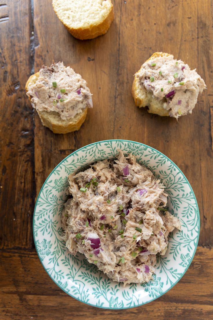 a bowl of tuna salad next to two pieces of bread on a wooden table top