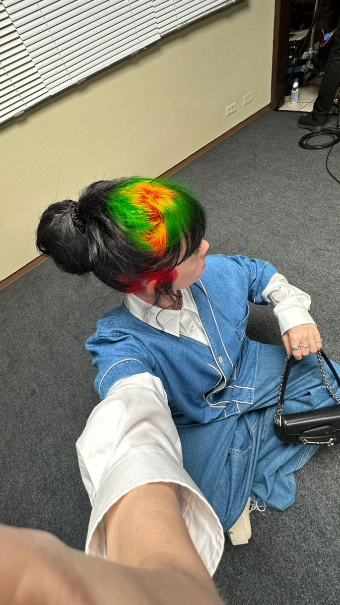 a person sitting on the floor with their hair dyed red and green, holding a black purse