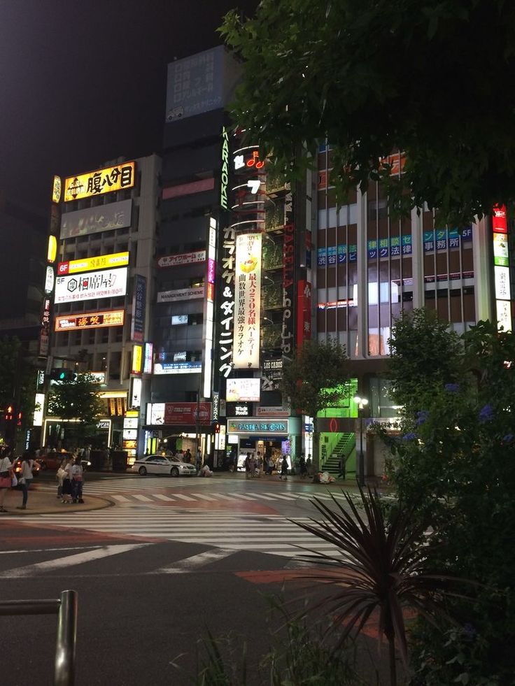 a city street at night with tall buildings in the background and people walking on the sidewalk