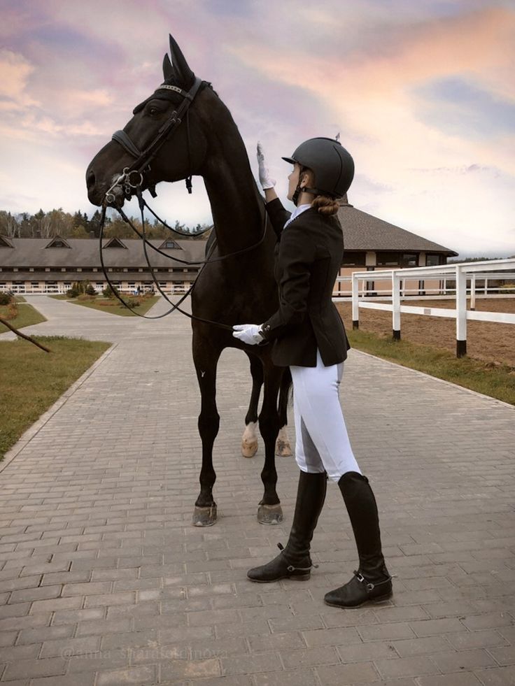 a woman in riding gear standing next to a horse on a brick walkway near a white fence