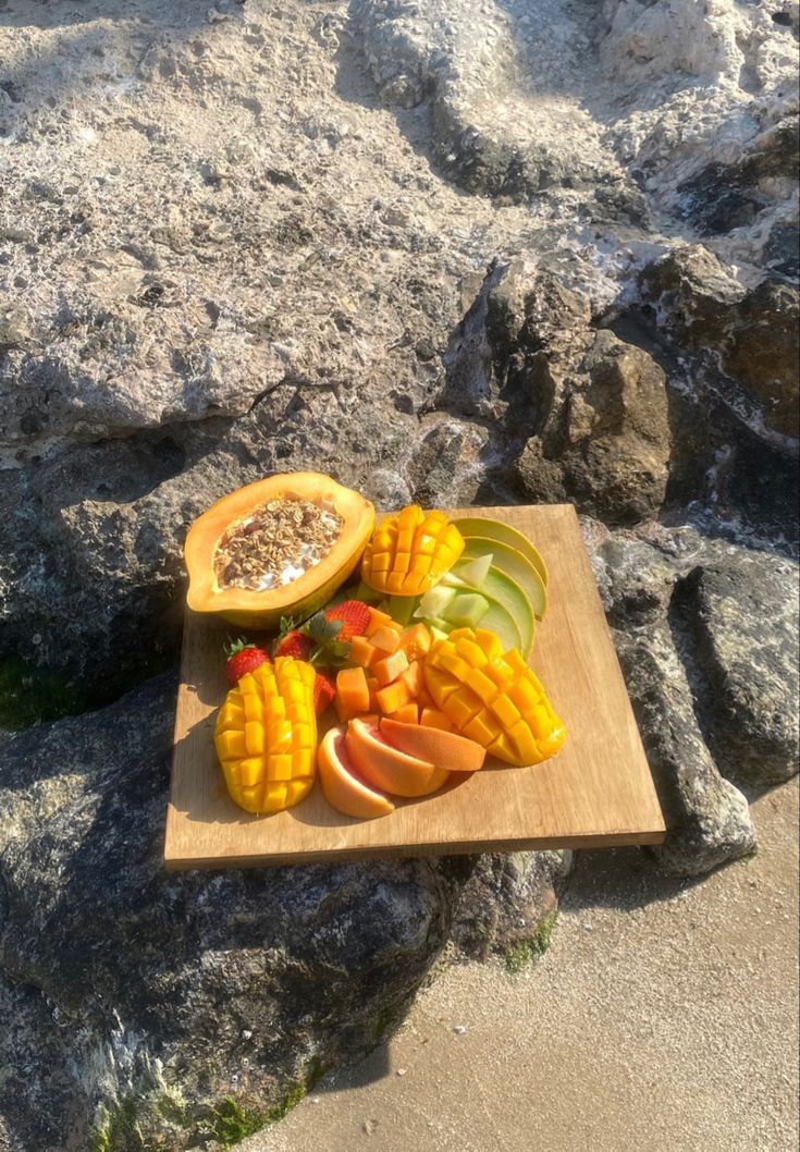 a wooden cutting board topped with fruit on top of a rock covered beach next to the ocean