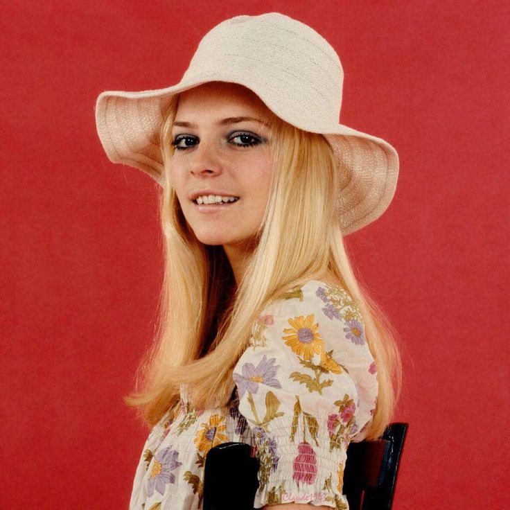 a woman with long blonde hair wearing a white hat and holding a black chair in front of a red background