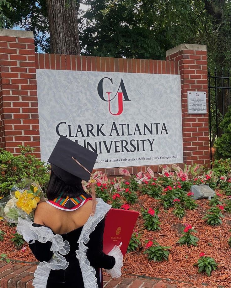 a woman in a graduation gown sitting on a bench outside clark atlanta university with flowers around her