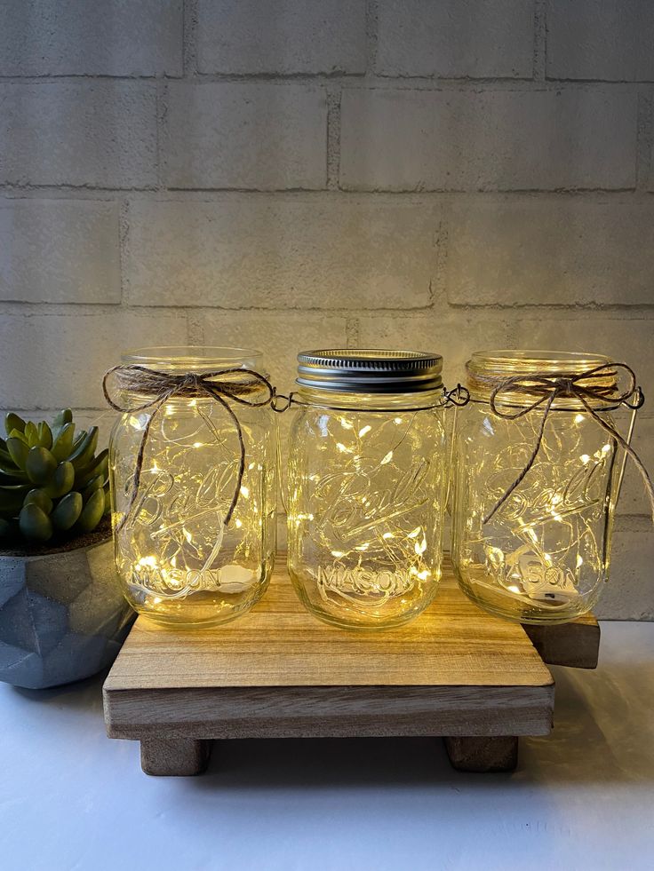 three mason jars with lights in them sitting on a wooden stand next to a succulent plant