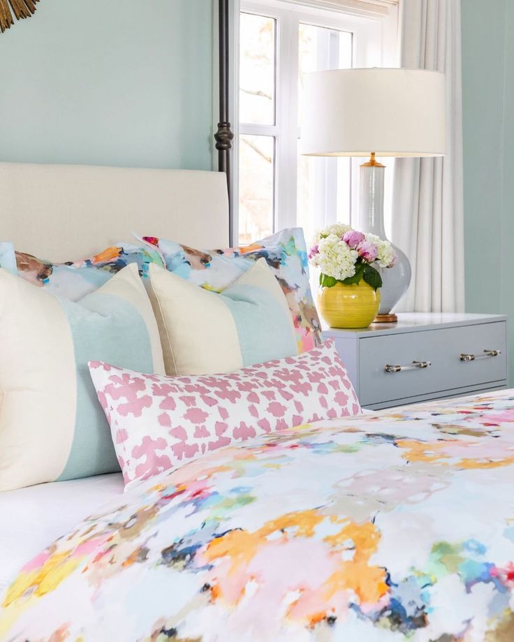 a bed with colorful sheets and pillows in a bedroom next to a dresser filled with flowers