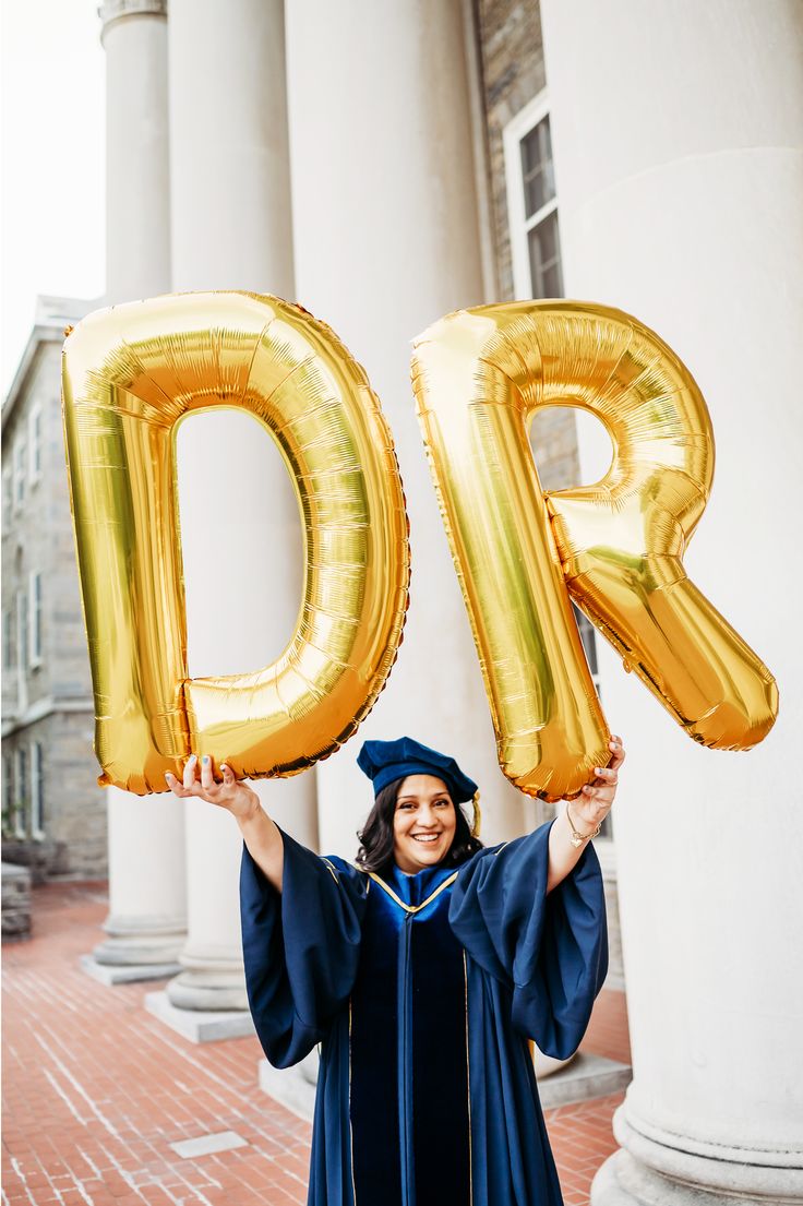 a woman in graduation gown holding up large gold balloons with the letters d and r