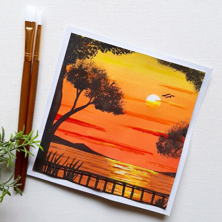 an orange sunset painting with two pencils next to it and a potted plant