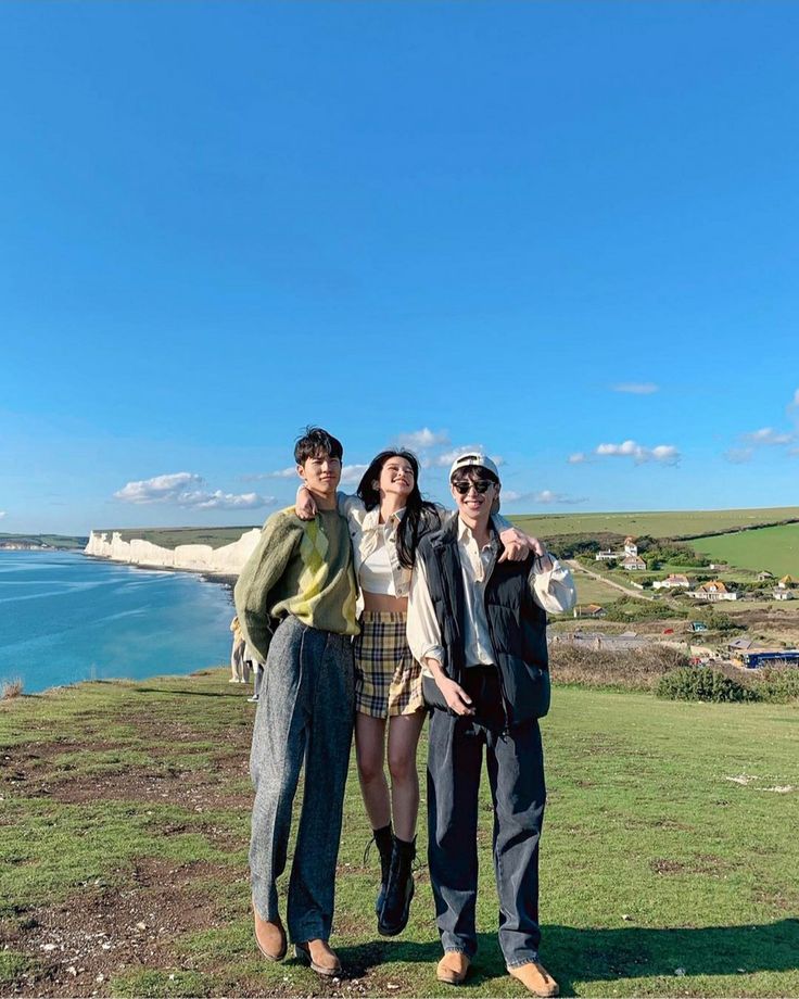 three people standing on top of a hill next to the ocean with their arms around each other