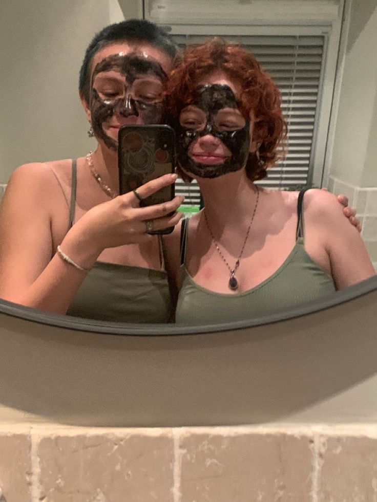 two women with masks on taking a selfie in front of a bathroom mirror while holding a cell phone