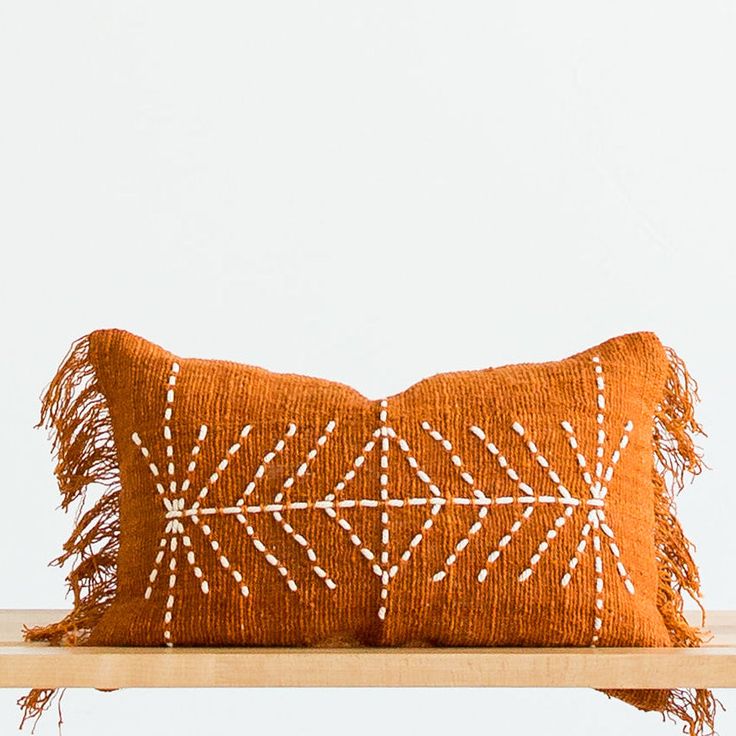 an orange pillow with white stitching on it sitting on a wooden shelf in front of a white wall