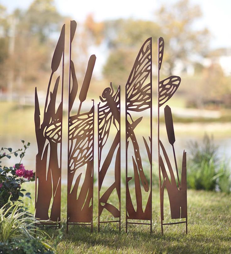 a metal sculpture sitting in the grass next to flowers and trees with dragonflies on them