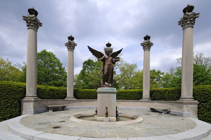 a statue in the middle of a circular area surrounded by pillars and bushes with statues on each side