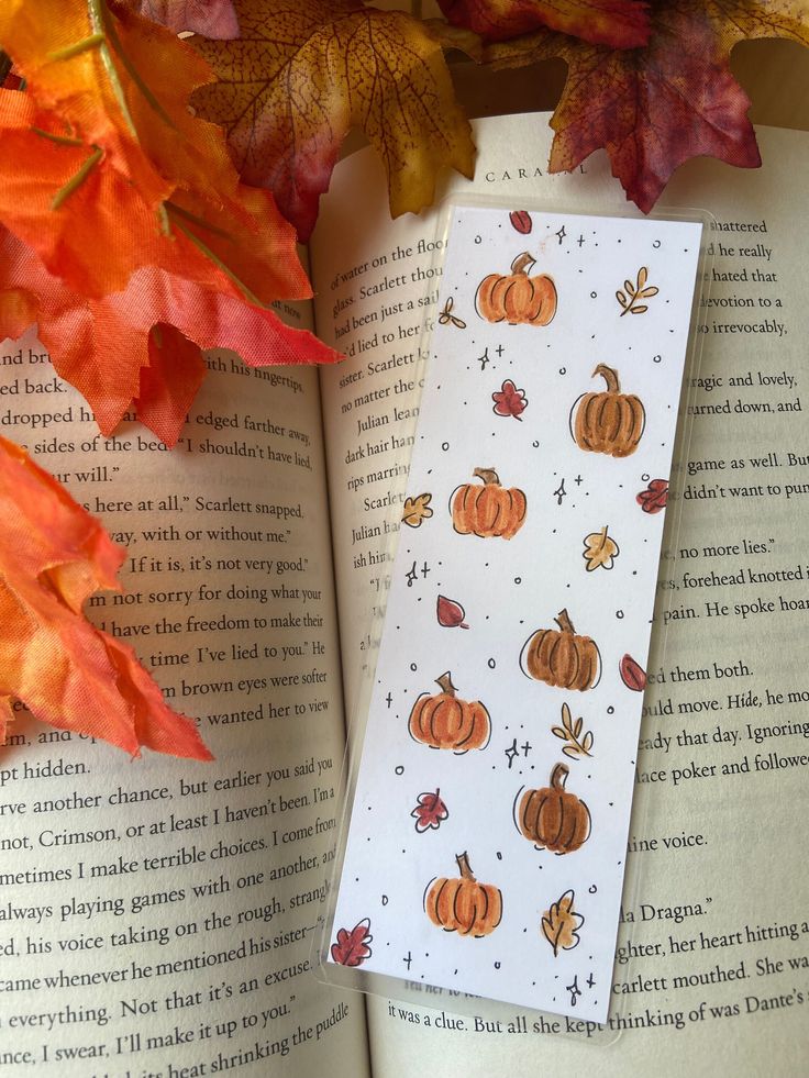 a bookmark with pumpkins and leaves on it sitting next to an open book