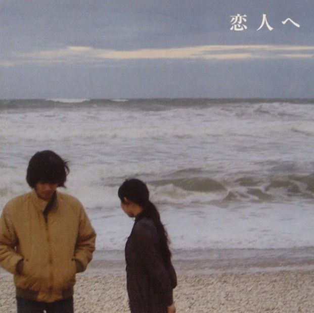 two people are standing on the beach looking at the waves in the water and one person is wearing a brown jacket