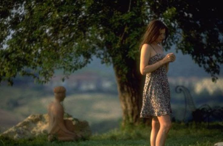 a woman standing in the grass looking at her cell phone while another person sits behind her