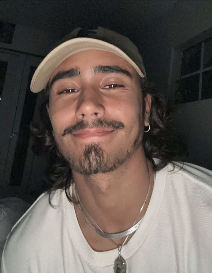 a man with long hair and a beard wearing a baseball cap smiles at the camera