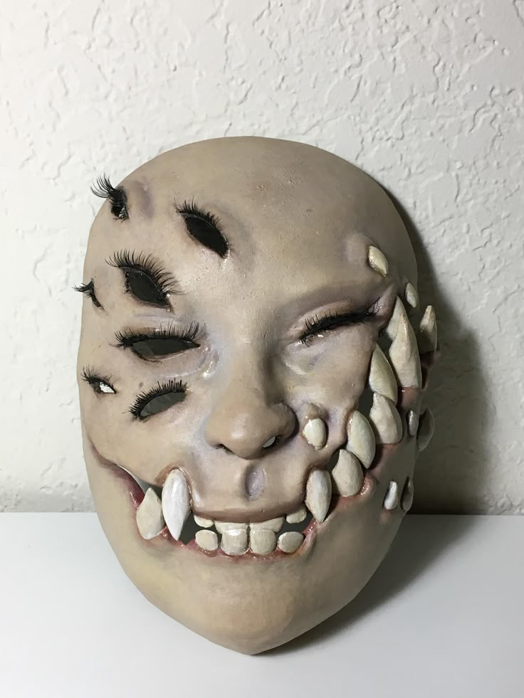 a white mask with black eyelashes and teeth on it's face, sitting against a wall