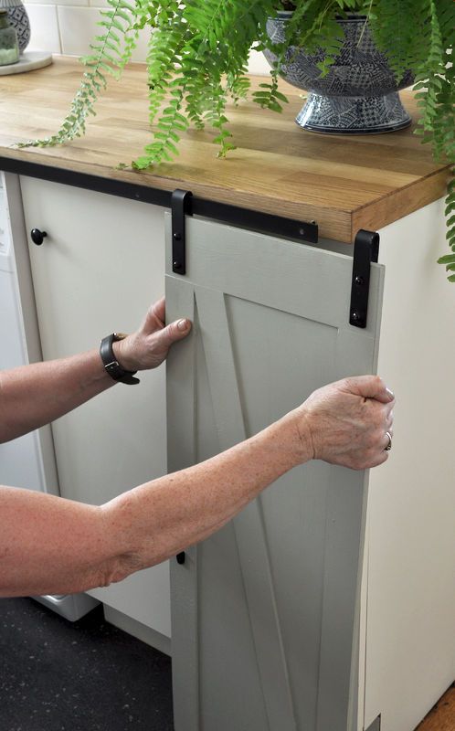 a woman is opening the door to a kitchen cabinet with plants on top of it