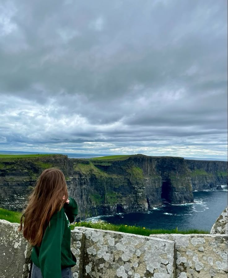 a woman standing on top of a stone wall next to the ocean looking at cliffs