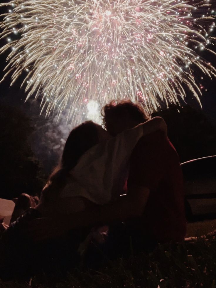 two people sitting on the ground with fireworks in the sky behind them and cars parked nearby