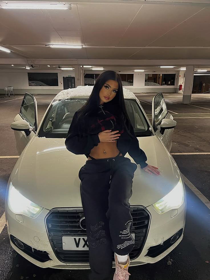 a woman sitting on top of a white car in a parking lot with her hands on her hips