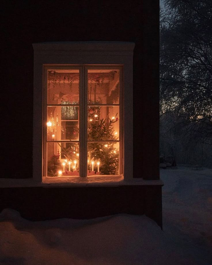 a lighted window with candles in the windowsill and snow on the ground around it