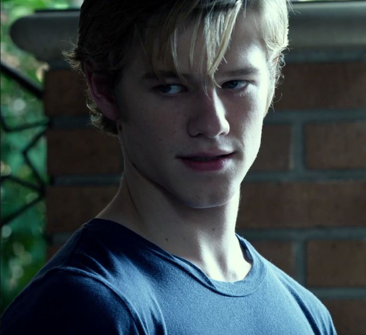 a young man with blonde hair standing in front of a brick wall and looking at the camera