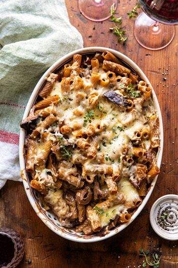 a casserole dish filled with pasta, mushrooms and parmesan cheese next to two glasses of wine