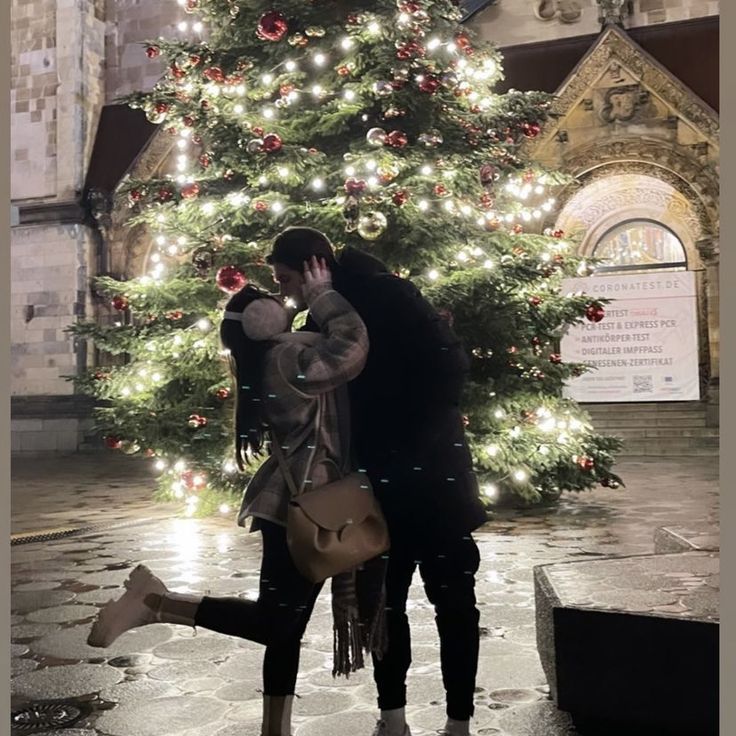 a man and woman kissing in front of a christmas tree with lights all around them