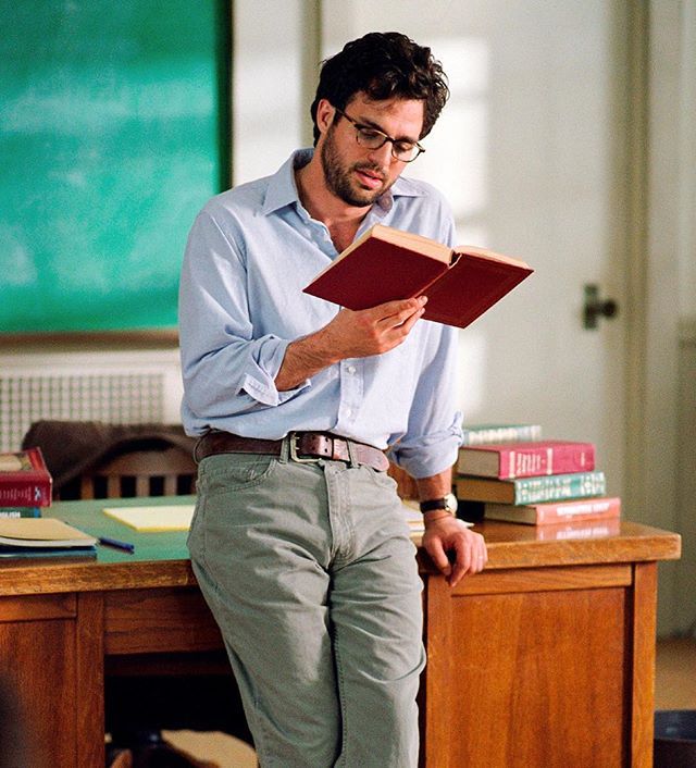 a man reading a book while standing in front of a desk with books on it