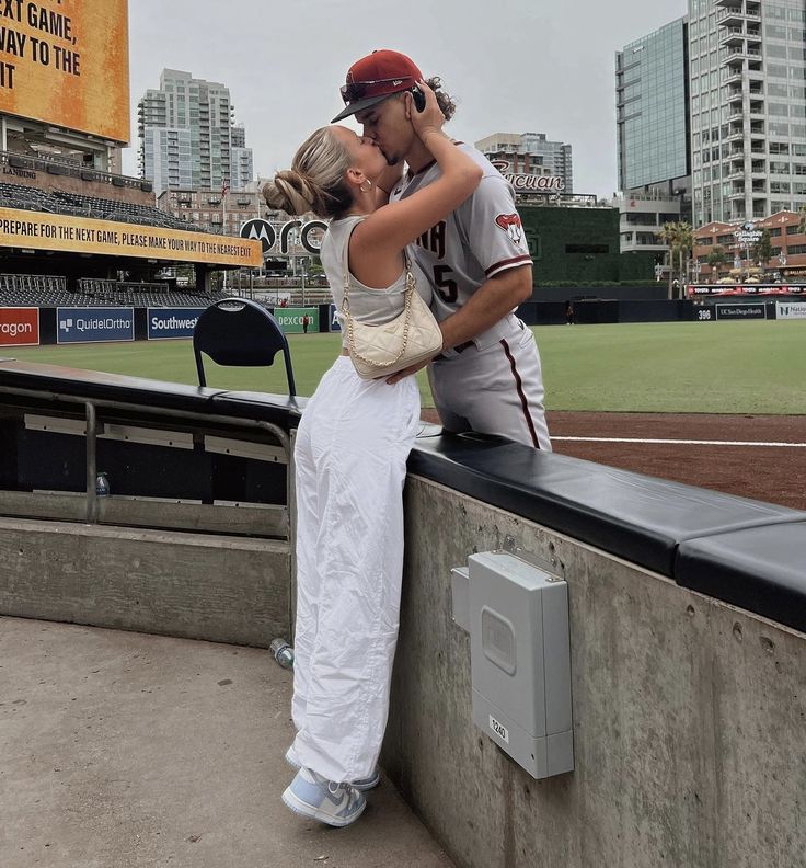 a man and woman kissing on the side of a baseball field with buildings in the background