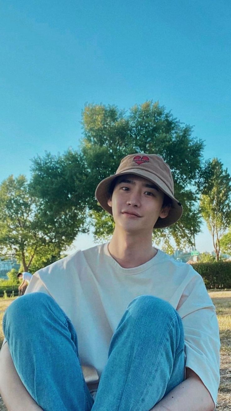 a young man sitting on the ground with his legs crossed wearing a hat and jeans