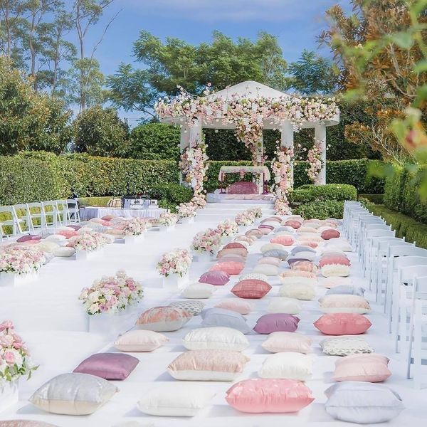 an outdoor wedding setup with pink and white pillows on the ground, surrounded by greenery