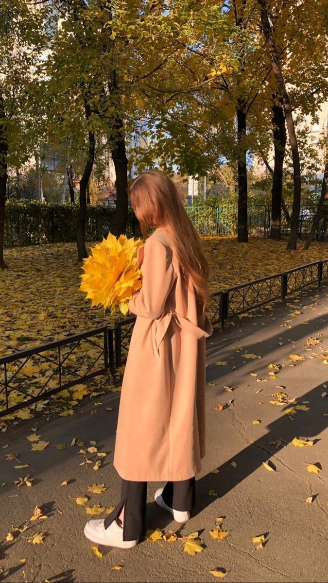 a woman in a trench coat is holding a sunflower and looking at the trees