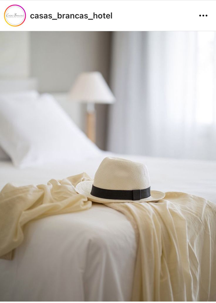 a white hat is laying on top of a bed with sheets and lamps in the background