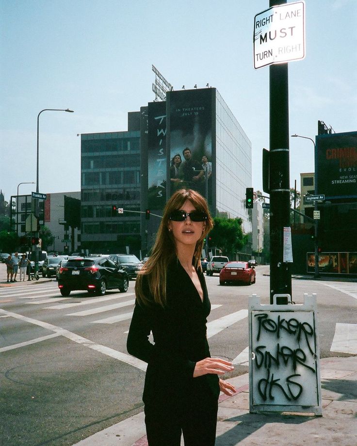 a woman standing on the side of a road next to a street sign with graffiti