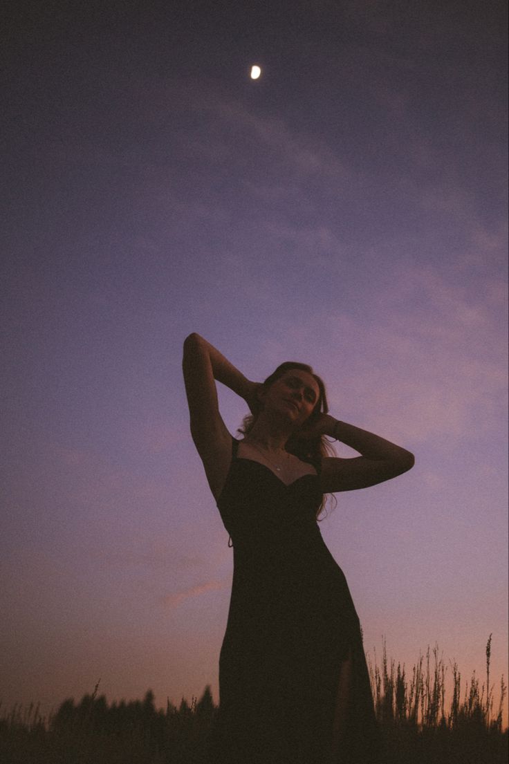 a woman in a long black dress is posing for the camera at sunset with her hand on her head